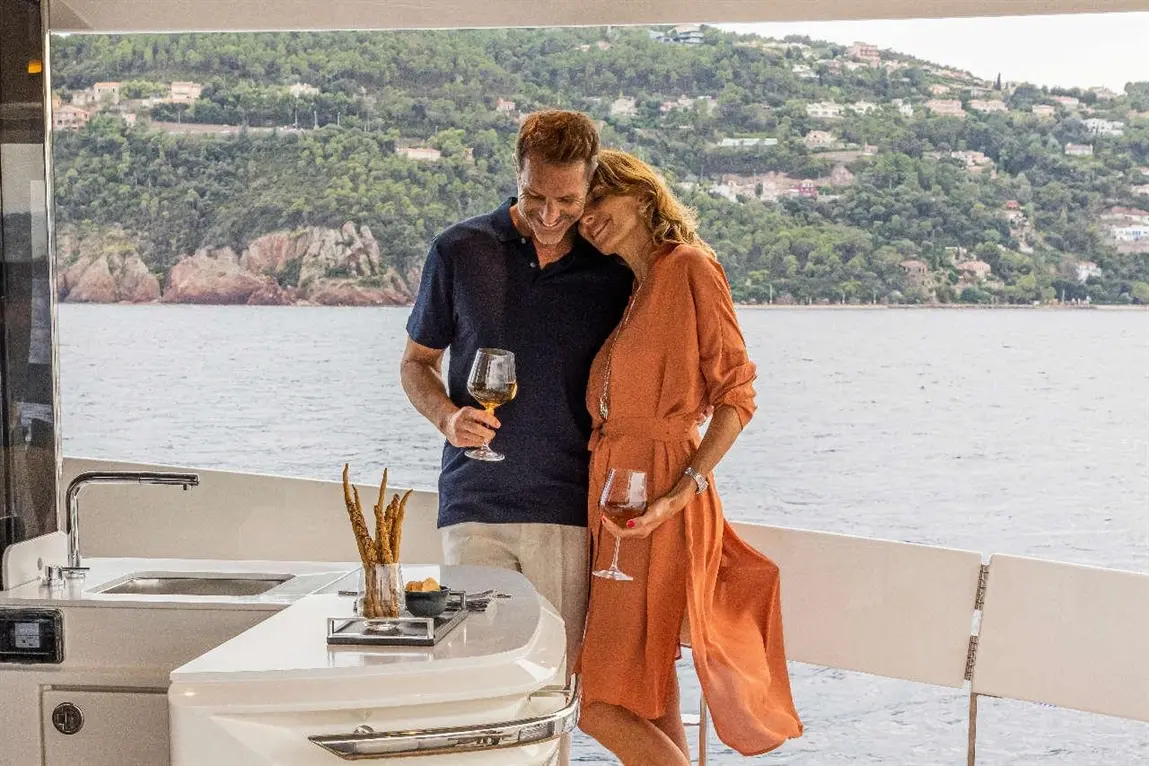 Couple enjoying drinks on a yacht, with a scenic coastal background and a stylish outdoor kitchen setup.